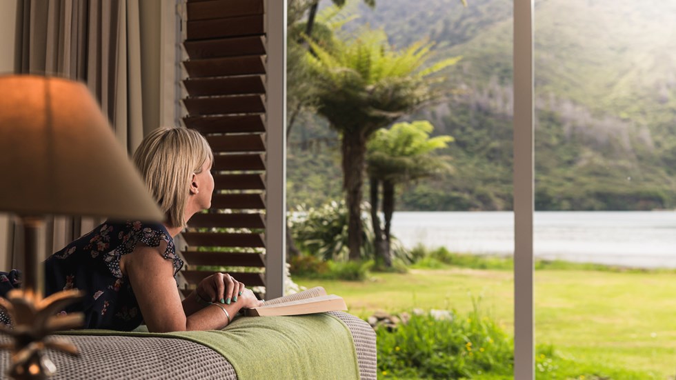 A lady enjoys the water view and a book from an Endeavour Suite at Furneaux Lodge in the Marlborough Sounds, New Zealand.