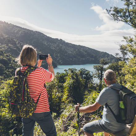 A woman takes a photo on her phone of the view across Ship Cove/Meretoto while a man sits on a bench on the northern Queen Charlotte Track in the Marlborough Sounds, New Zealand.