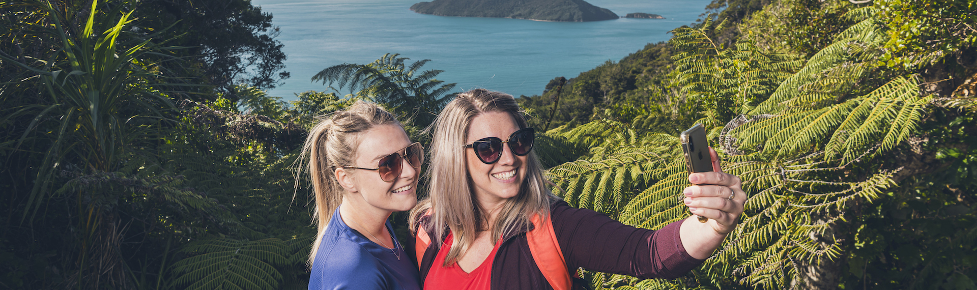 Two women hikers take a selfie photo along Queen Charlotte Track in the Marlborough Sounds, New Zealand, with Motuara Island and sea view in distance.