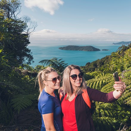 Two women hikers take a selfie photo along Queen Charlotte Track in the Marlborough Sounds, New Zealand, with Motuara Island and sea view in distance.