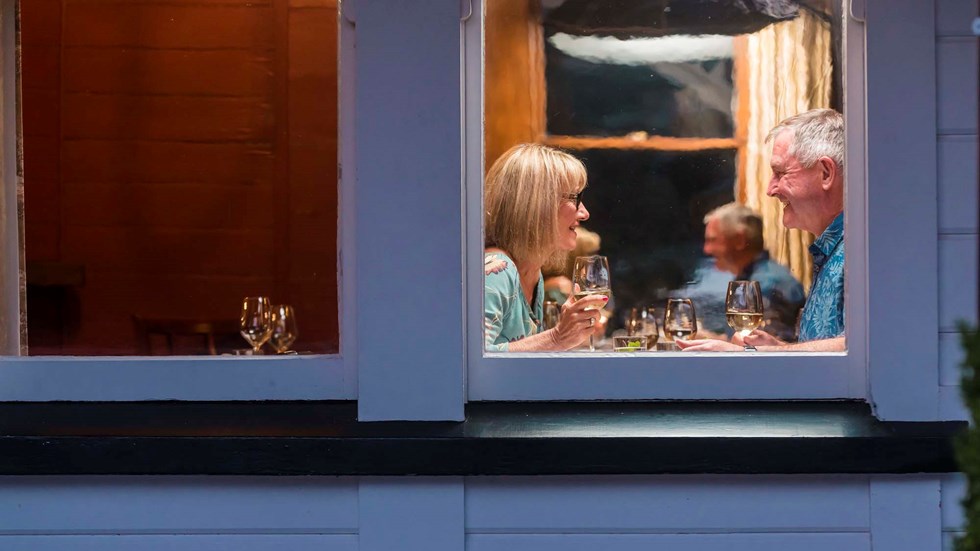 A couple enjoy glasses of local Marlborough wine at a window table during dinner at Furneaux Lodge in the Marlborough Sounds, New Zealand.